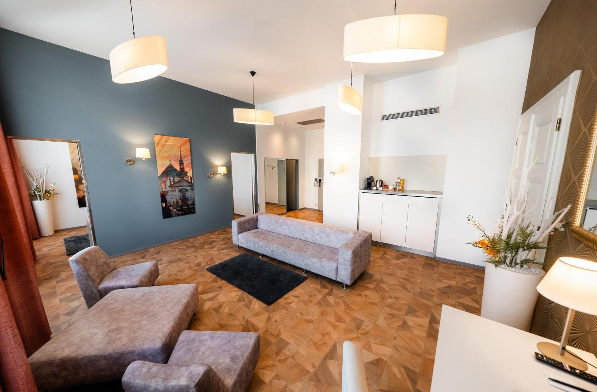 Grandium Hotel Prague Exterior photo The photo shows a modern and stylish living space. The room features light-colored walls and a distinct blue accent wall. There is a large, gray sectional sofa and a small black rug on the wooden floor. The space is well-lit with two pendant lamps ha