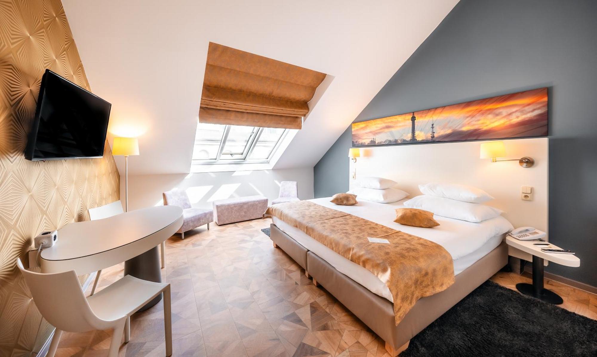 Grandium Hotel Prague Exterior photo The photo shows a stylish and modern hotel room with an angled ceiling. It features a large bed with white linens and decorative pillows at the center. A soft, fur-like throw covers part of the bed. There are two bedside lamps and a wall-mounted TV o